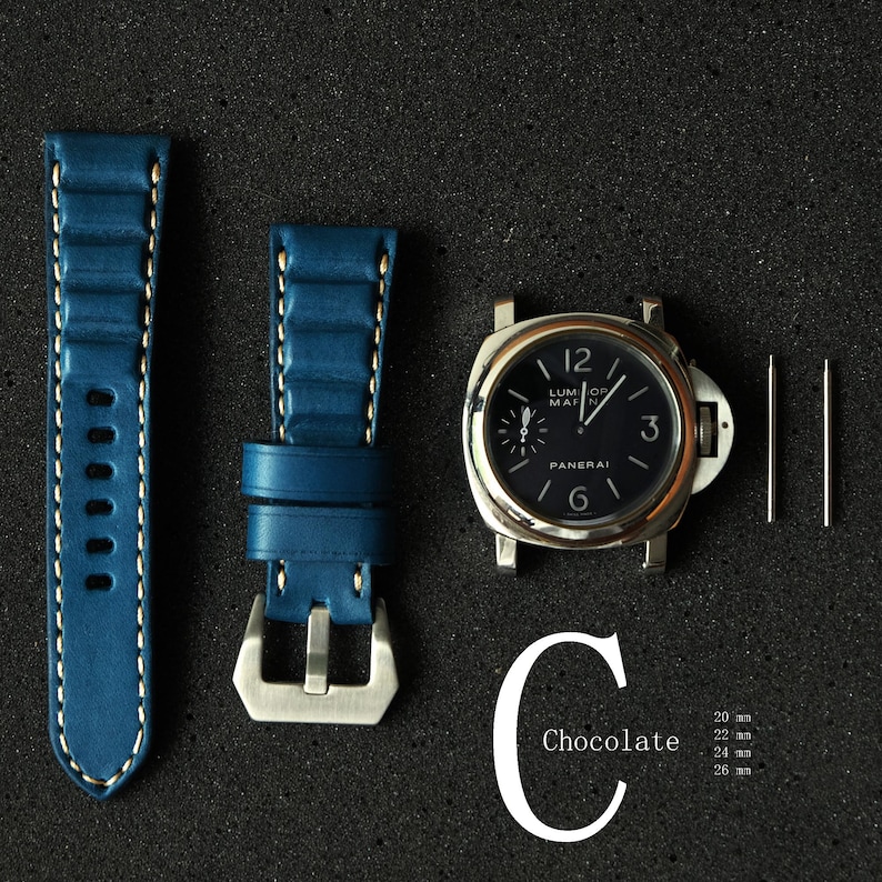 May include: A watch face with a black dial and silver casing, accompanied by a blue leather watch strap with white stitching and a silver buckle. Two silver pins are also included. The word "Chocolate" is written in white.