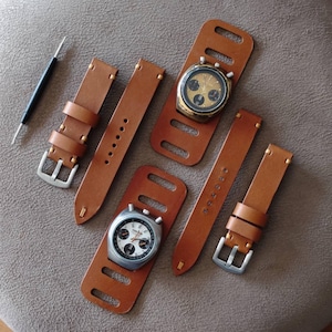 May include: A collection of brown leather watch straps and two vintage chronographs. The straps have silver buckles and are of varying lengths. One watch has a gold-toned face, the other a silver-toned face. A watch tool is also present.