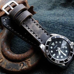 May include: A black Seiko watch with a silver bezel and a black leather strap. The watch has a black dial with white markers and hands. The strap is gray and stitched with white thread. The watch is resting on a rusty metal object.