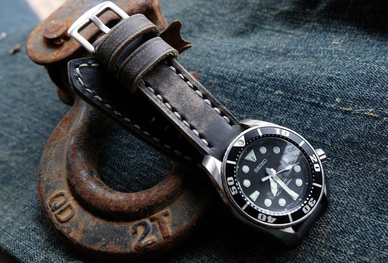 May include: A black Seiko watch with a silver bezel and a black leather strap. The watch has a black dial with white markers and hands. The strap is gray and stitched with white thread. The watch is resting on a rusty metal object.
