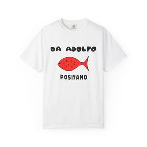 May include: White t-shirt with the text "DA ADOLFO POSITANO" in black, above a red fish illustration. The shirt is a casual style, likely made of cotton, with a crew neck.