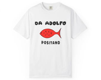 以下が含まれることがあります： 白いTシャツ。黒い文字で「DA ADOLFO POSITANO」と書かれ、赤い魚のイラストが描かれています。カジュアルなスタイルで、綿素材のクルーネックTシャツです。
