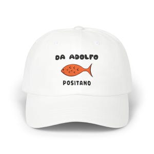May include: White baseball cap with the text "DA ADOLFO POSITANO" above a cartoon orange fish. The cap is made of a soft material and has a curved brim.