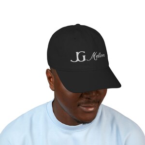 May include: Black baseball cap with a curved peak. The cap has the embroidered text "JG Melon" in a cursive font. The cap is worn by a person wearing a light blue top.