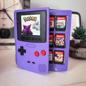 May include: A purple plastic game cartridge storage case shaped like a handheld console. The front features a screen displaying Pokémon graphics and the text GAMES BOOK. The interior holds six Nintendo Switch game cartridges in individual slots.