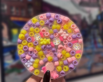 May include: A round, clear resin coaster filled with colorful beads in shades of yellow, purple, and pink. The coaster also features small, decorative candies and a swirl design.