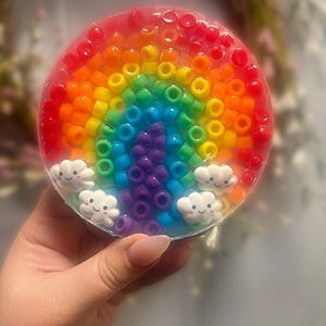 May include: A round, white soap with a rainbow made of colourful beads and three white clouds with happy faces.