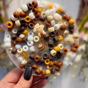 Op de afbeelding: Een ronde onderzetter van doorschijnende hars, ingelegd met een verscheidenheid aan kleine, ronde plastic kralen in tinten bruin, beige, wit en geel. Het ontwerp bevat twee kleine, decoratieve figuurbedeltjes: een crèmekleurige hond en een bruine beer.