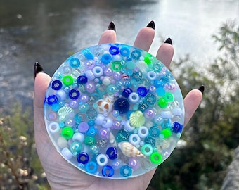 May include: A clear round resin coaster with embedded blue, green, white, and iridescent beads, seashells, and other small decorative elements.