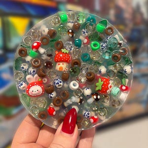 Puede incluir: Un posavasos redondo de resina transparente lleno de cuentas de colores y elementos decorativos en miniatura. El posavasos presenta una fresa, una seta y otros pequeños detalles caprichosos. El posavasos tiene aproximadamente 10 cm de diámetro.