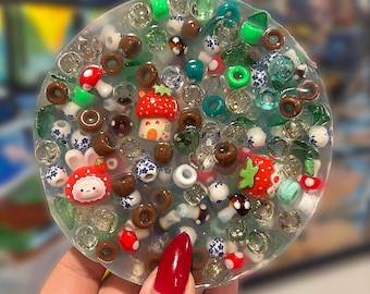 May include: A circular clear resin coaster embedded with a variety of colorful beads, including small brown rings, green translucent shapes, white floral patterned spheres, and miniature red strawberry-themed charms and mushroom figures.