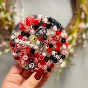 May include: A clear, round resin coaster filled with red, white, and black beads, small skulls, and a razor blade. The coaster has a spooky, Halloween theme with a variety of decorative elements.