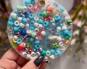 May include: A circular resin coaster embedded with a variety of colorful beads and charms. The design features numerous blue, white, and green plastic beads mixed with small sea-themed figures including starfish, seashells, and a small red fish.