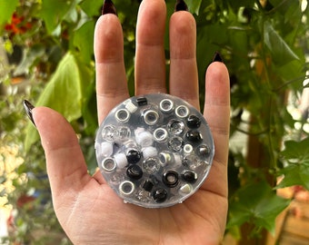 May include: A clear resin tray filled with black, white, and silver beads. The tray is being held in a person's hand.