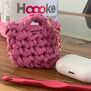 Op de afbeelding: Een roze gehaakte tas met een gouden sleutelhanger en een witte AirPod-hoes. Een roze 8,0 mm haaknaald is ook afgebeeld.