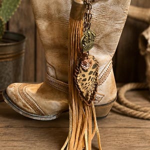 May include: A light tan leather boot featuring a distressed finish, decorative stitching, and a buckle strap. Attached to the side is a long tan suede tassel charm with a metal chain, a small crown pendant, and a leopard print leather leaf accent.