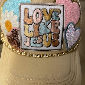 May include: Tan trucker hat with embroidered patches of a daisy, hearts, and a square patch with the words "LOVE LIKE JESUS". The hat features a gold chain with pearls along the brim.