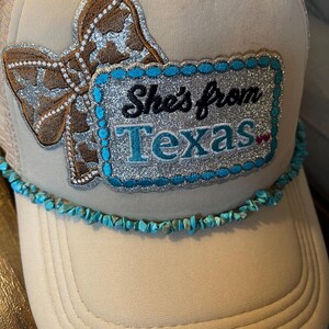 May include: Tan trucker hat with a brown and silver bow applique and a turquoise stone trim. The hat features a patch that reads "She's from Texas" in black and turquoise lettering.