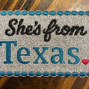 May include: A rectangular patch with a silver glitter background and a teal scalloped border. The text "She's from Texas" is embroidered in black and teal, with two small red hearts at the end.