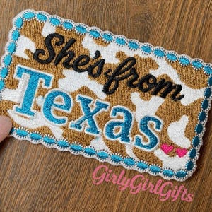 May include: Embroidered patch with the text "She's from Texas" in black and turquoise. The background has a cowhide pattern in brown and white, with a turquoise border. The patch also includes pink hearts and the text "GirlyGirlGifts".