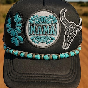 May include: Black trucker hat with turquoise and silver accents. Features a patch with the word "MAMA" in turquoise, a cactus, and a steer skull. The brim is adorned with turquoise beads.