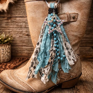 May include: A tan leather cowboy boot with a decorative star-shaped charm. The charm features blue beads, a silver star with purple accents, and flowing fabric in blue and animal print. The boot is on a wooden surface.
