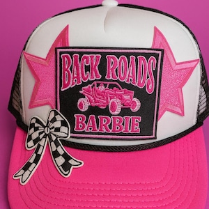 May include: A trucker hat with a pink brim and a white front panel featuring a black rectangle with the words "BACK ROADS BARBIE" and a pink off-road vehicle graphic. Pink star accents and a checkered bow complete the design.