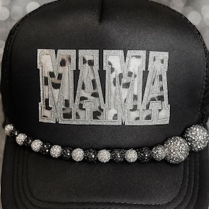 May include: Black trucker hat with the word "MAMA" in silver glitter letters with a cow print design. The hat has a beaded embellishment along the brim. A stylish accessory.