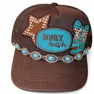 May include: Brown trucker hat with a mesh back and a curved brim. The front features a star and boot applique, a turquoise oval with "HONEY hush" text, and a decorative band with turquoise accents. A stylish accessory.