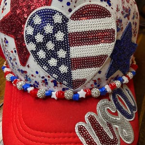 May include: A white baseball cap featuring a large sequined heart with an American flag pattern, a red sequined star, and a blue sequined star. The cap is adorned with red, white, and blue beads and the word USA in silver and blue sequins.