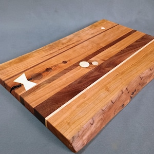 May include: A rectangular wooden cutting board with a natural edge. The board features a mix of light and dark wood tones, with inlaid accents including a bow tie shape and circular designs. The board is approximately 18 inches long and 12 inches wide.