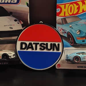 May include: A round Datsun logo in red, white, and blue, with the word "Datsun" in white block letters. The logo is set against a dark background, with toy cars in the background.