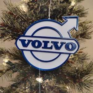 May include: A white and blue 3D printed Christmas ornament featuring the Volvo logo. The ornament is shaped like the Volvo logo with a blue arrow pointing to the right.