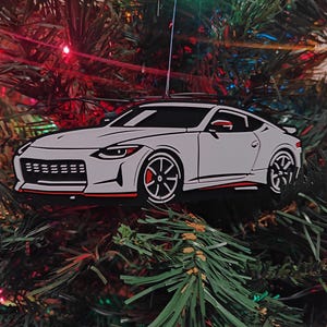 May include: A white car-shaped Christmas tree ornament with black wheels and red detailing. The car is suspended amongst the green needles of a Christmas tree. The ornament is a festive decoration for car lovers.