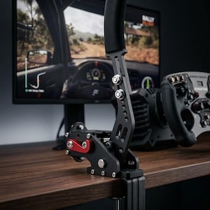 May include: Black and silver racing simulator handbrake with a black padded handle and red accents. The handbrake is mounted to a desk with a clamp. A racing wheel and monitor displaying a racing game are in the background.