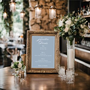 K&ouml;nnte beinhalten: Ein goldfarbener Rahmen mit einem hellblauen Hintergrund und wei&szlig;em Text, der "Signature Drinks" lautet. Das Schild listet eine Vielzahl von Getr&auml;nken mit Namen wie "One and Two", "Here's to", "Cheers" und "Bailey's & Brandy" auf.