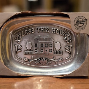 Può includere: Piatto in metallo color argento con le parole "BLESS THIS HOUSE" e un disegno di una casa. Il piatto è in una scatola di cartone con le parole "AMERICAN INSPIRATIONS" sul lato. Il piatto ha un design floreale e a cuore decorativo.