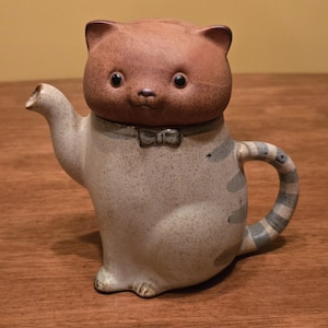 Op de afbeelding: Een keramische theepot in de vorm van een kat. De katvormige theepot heeft een bruine kop en een lichtgrijze body met blauwe strepen op het handvat. Het heeft een kleine strik en een tuit.