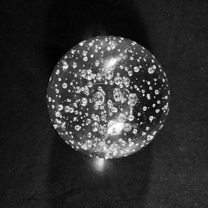 May include: A clear glass sphere with many small air bubbles trapped inside. The sphere is reflecting light and casting a shadow on a black surface.