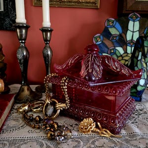 May include: A collection of antique items including a red decorative box, a gold necklace, and a gold flower brooch. Two silver candlesticks with white candles are behind the box. A stained glass ornament is in the background. The box is approximately 15 cm long.