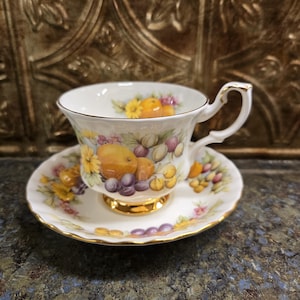 以下が含まれることがあります： フルーツ柄の白いティーカップとソーサーのセット。 ティーカップとソーサーには金のトリムとハンドルが付いています。 フルーツのデザインには、オレンジ、ブドウ、黄色の花が含まれています。 セットは暗い表面にあります。