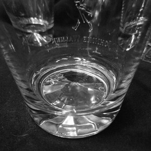 May include: Clear, faceted glass tumbler with the "Johnnie Walker" logo etched on the side. The glass has a thick base and a slightly flared shape. The image is in black and white.