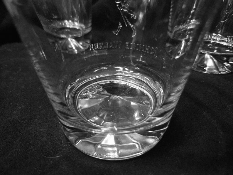 May include: Clear, faceted glass tumbler with the "Johnnie Walker" logo etched on the side. The glass has a thick base and a slightly flared shape. The image is in black and white.