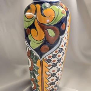 May include: A ceramic vase featuring a hand-painted design with a dark blue rim and neck. The body displays alternating panels of vibrant orange, green, and brown swirling patterns alongside a white grid section with small floral accents.