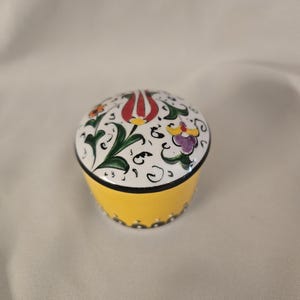 Puede incluir: Pequeña caja redonda de cerámica con tapa. La caja es amarilla con un borde negro y una tapa blanca decorada con diseños florales rojos, morados y naranjas. La tapa tiene un borde negro.
