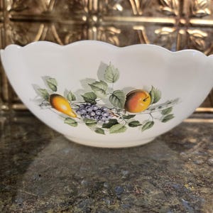 May include: A white ceramic bowl with a decorative, scalloped edge. The bowl features a fruit design with a pear, grapes, and a peach, along with green leaves. The bowl is suitable for serving.