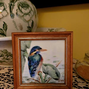 May include: A framed watercolour painting of a kingfisher bird perched on a branch. The bird has blue, green, and brown feathers, with a long orange beak. The painting is in a square wooden frame with a gold inner border. The background shows water and lily pads.