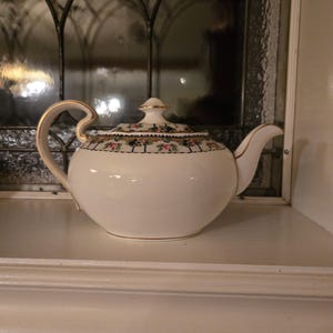 May include: A white porcelain teapot with a gold handle and trim. The lid features a floral pattern with pink and green accents. The teapot is displayed on a white shelf in front of a leaded glass window.