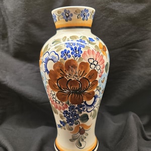 May include: A cream-colored ceramic vase with a flared top and base, adorned with hand-painted floral designs. The vase features a large brown flower as the focal point, surrounded by blue, pink, and orange blossoms. The rim and base are accented with orange and blue bands.