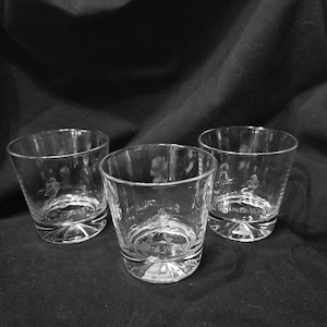 May include: Three clear, lowball whiskey glasses with a rounded base and a slightly flared rim. Each glass has an etched design and the text "JOHNNIE WALKER" on the base. The glasses are arranged on a dark surface.